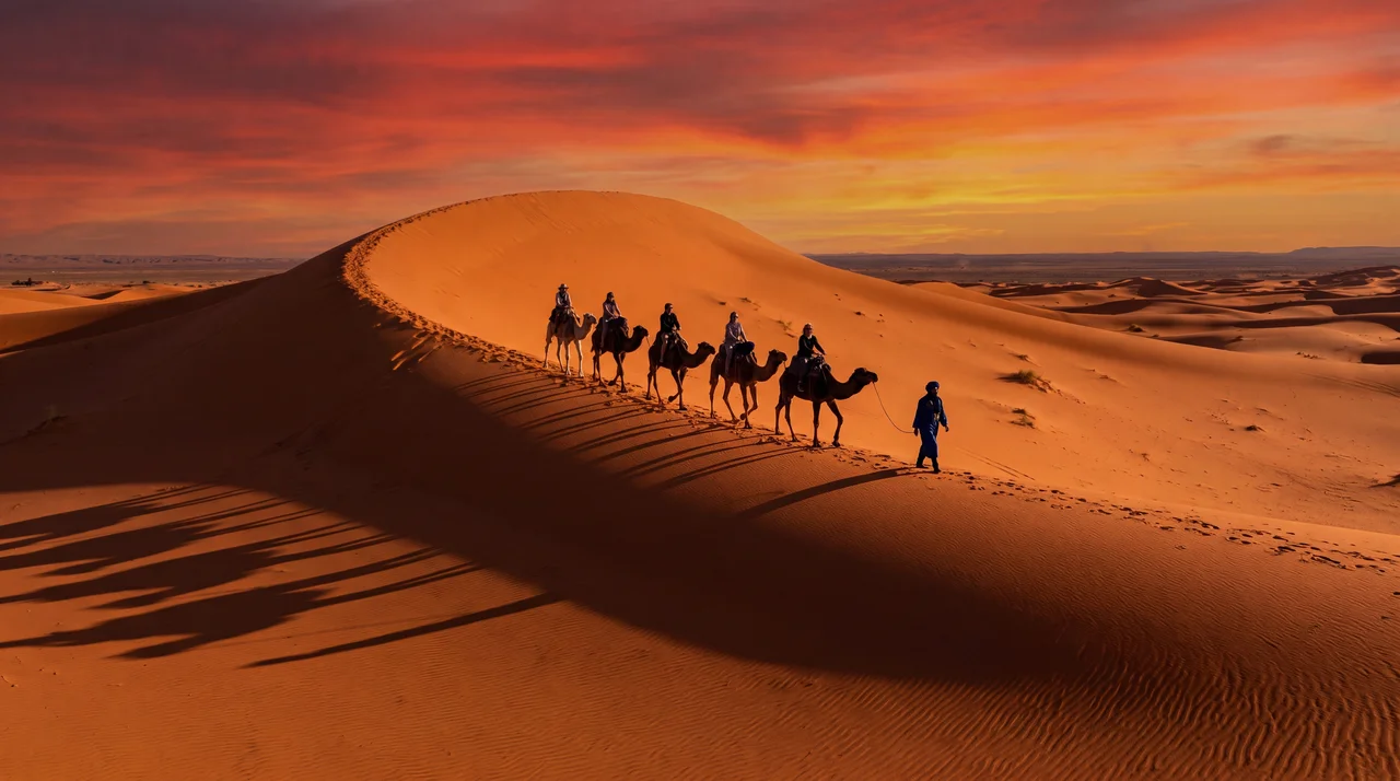 Camel riding Morocco — a line of five camels with riders and Berber guide walking along the crest of Erg Chebbi sand dunes at sunset near Merzouga, Sahara Desert Morocco 2026